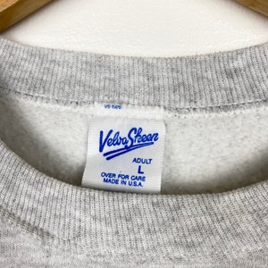 K&ouml;nnte beinhalten: Grauer Sweatshirt mit einem blauen Velva Sheen-Etikett. Das Etikett zeigt "Adult" und "L" f&uuml;r die Gr&ouml;&szlig;e. Au&szlig;erdem steht "Over for care" und "Made in U.S.A."