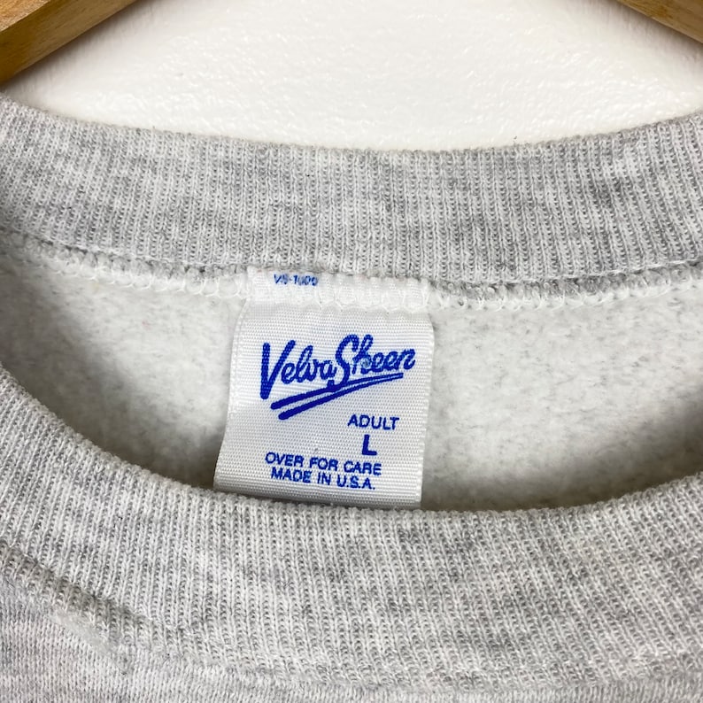 K&ouml;nnte beinhalten: Grauer Sweatshirt mit einem blauen Velva Sheen-Etikett. Das Etikett zeigt "Adult" und "L" f&uuml;r die Gr&ouml;&szlig;e. Au&szlig;erdem steht "Over for care" und "Made in U.S.A."