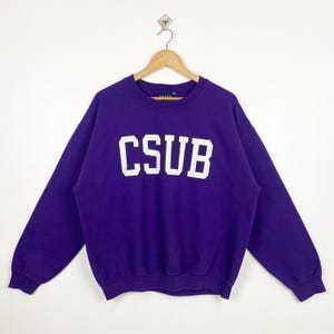 May include: A purple sweatshirt with the white letters "CSUB" across the chest. The sweatshirt is hanging on a wooden hanger against a white background. The sweatshirt has long sleeves and a crew neck.