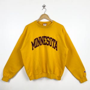 May include: A yellow sweatshirt with a maroon "MINNESOTA" arch logo on the front. The sweatshirt has a Champion logo on the left sleeve.