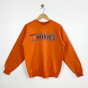 Può includere: Felpa Champion arancione con la scritta "HOKIES" in bianco e nero, con "VIRGINIA TECH" sopra. La felpa ha maniche lunghe e girocollo. Il logo Champion è sulla manica sinistra.