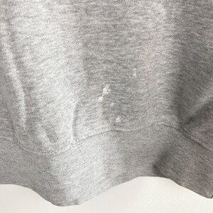 May include: Close-up of a heather gray sweatshirt with white paint splatters. The image shows the bottom hem and a portion of the body of the garment. The fabric has a textured appearance.