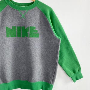 Pu&ograve; includere: Felpa grigia con maniche e finiture verdi. La parola "NIKE" &egrave; stampata a grandi lettere verdi sul petto. La felpa ha un girocollo e maniche lunghe.