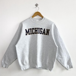 Peut inclure: Un sweat-shirt à col rond gris chiné clair avec le mot MICHIGAN imprimé en lettres capitales bleu marine foncé, soulignées d'un fin contour jaune sur la poitrine. Le vêtement présente des manches longues, un col, des poignets et un ourlet côtelés.