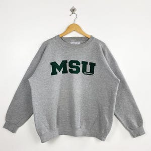 Puede incluir: Sudadera gris con las letras "MSU" en verde oscuro en el pecho. La palabra "Spartans" está impresa debajo. La sudadera está colgada de una percha de madera.
