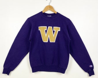 Op de afbeelding: Paarse trui met een grote gouden "W" op de voorkant. De trui heeft lange mouwen en een ronde hals. Het Champion-logo bevindt zich op de linkermouw.