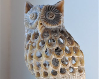 May include: A carved owl figurine made of light brown stone. The owl is perched on a branch with a detailed base. The owl's eyes are large and round, and its feathers are intricately carved.