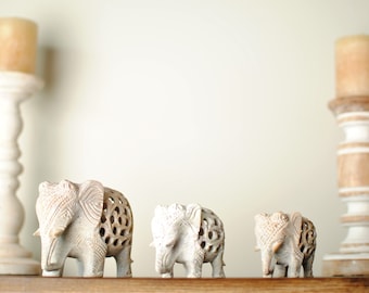 May include: Three white carved elephant figurines on a wooden shelf with two white candle holders on either side.