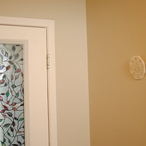 May include: A white door with a stained glass window featuring a leafy design in shades of green, blue, purple, and red. The door is set against a wall painted in a light beige color. A white octagonal wall decor with a floral design is mounted on the adjacent wall, which is painted in a light brown color.