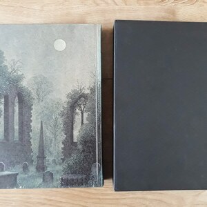 K&ouml;nnte beinhalten: Ein Hardcover-Buch mit einem grauen und wei&szlig;en Einband, der einen mondbeschienenen Friedhof mit einem Steinbogen und B&auml;umen zeigt. Das Buch ist geschlossen und hat einen schwarzen Schutzumschlag.