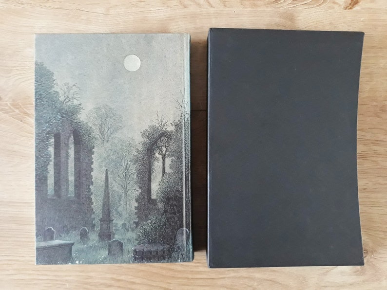K&ouml;nnte beinhalten: Ein Hardcover-Buch mit einem grauen und wei&szlig;en Einband, der einen mondbeschienenen Friedhof mit einem Steinbogen und B&auml;umen zeigt. Das Buch ist geschlossen und hat einen schwarzen Schutzumschlag.