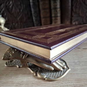 Pu&ograve; includere: Un libro rilegato in pelle bordeaux con dettagli dorati e un bordo dorato decorativo. Il libro &egrave; appoggiato su un fermacarte in ottone a forma di mano.