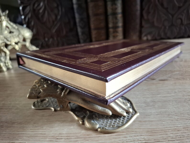 Pu&ograve; includere: Un libro rilegato in pelle bordeaux con dettagli dorati e un bordo dorato decorativo. Il libro &egrave; appoggiato su un fermacarte in ottone a forma di mano.
