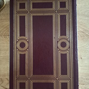 Pu&ograve; includere: Un libro rilegato in pelle bordeaux con un design decorativo dorato sulla copertina. Il design presenta una serie di cerchi concentrici e quadrati.