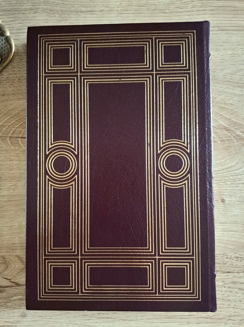 Pu&ograve; includere: Un libro rilegato in pelle bordeaux con un design decorativo dorato sulla copertina. Il design presenta una serie di cerchi concentrici e quadrati.