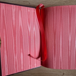 Pu&ograve; includere: Un segnalibro a nastro rosso &egrave; attaccato a un libro chiuso con una fodera in raso rosa. Il libro &egrave; aperto per rivelare le pagine bianche.