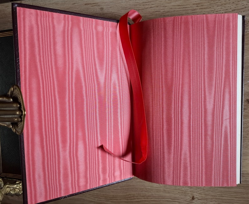 Pu&ograve; includere: Un segnalibro a nastro rosso &egrave; attaccato a un libro chiuso con una fodera in raso rosa. Il libro &egrave; aperto per rivelare le pagine bianche.