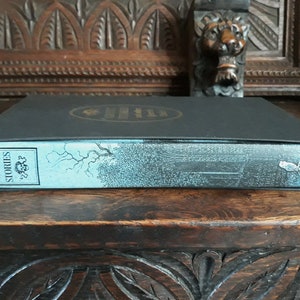 K&ouml;nnte beinhalten: Ein schwarzes Hardcover-Buch mit einem blauen R&uuml;cken. Der R&uuml;cken tr&auml;gt den Titel "Ghost Stories" und das Wort "Folio" ist darauf gedruckt. Das Buch liegt auf einer Holzoberfl&auml;che.