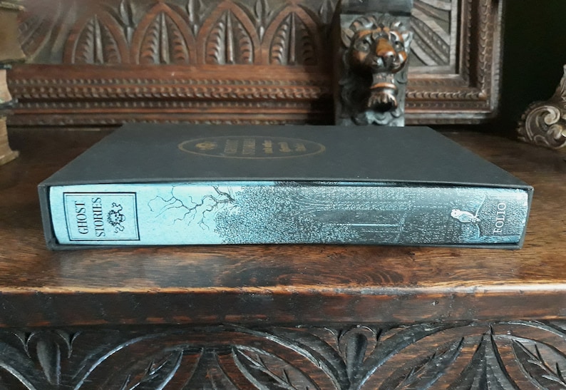 K&ouml;nnte beinhalten: Ein schwarzes Hardcover-Buch mit einem blauen R&uuml;cken. Der R&uuml;cken tr&auml;gt den Titel "Ghost Stories" und das Wort "Folio" ist darauf gedruckt. Das Buch liegt auf einer Holzoberfl&auml;che.