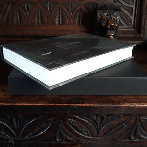 K&ouml;nnte beinhalten: Ein schwarzes Hardcover-Buch mit wei&szlig;em R&uuml;cken und einem schwarzen Schuber. Das Buch liegt auf einer Holzoberfl&auml;che.