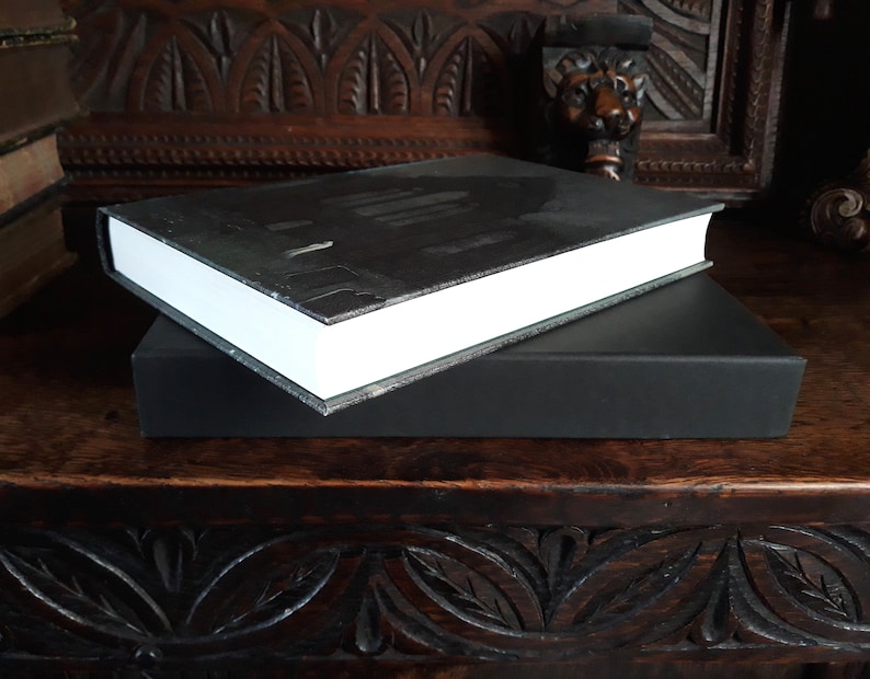K&ouml;nnte beinhalten: Ein schwarzes Hardcover-Buch mit wei&szlig;em R&uuml;cken und einem schwarzen Schuber. Das Buch liegt auf einer Holzoberfl&auml;che.