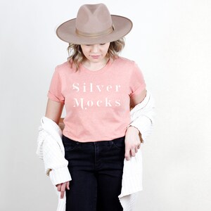 Op de afbeelding: Een vrouw draagt een perzikkleurig T-shirt met de tekst "Silver Mocks" erop gedrukt. Ze draagt een witte cardigan en een bruine fedora.