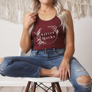 Può includere: Un top a canotta color granata con un designo floreale bianco e il testo "Silver Mocks" sul davanti. La modella indossa jeans blu con dettagli consumati.