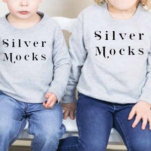 K&ouml;nnte beinhalten: Zwei Kinder tragen graue Langarmshirts mit dem Aufdruck "Silver Mocks" auf der Vorderseite. Beide tragen blaue Jeans und sitzen auf einem wei&szlig;en Stuhl.