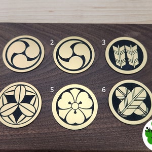 Puede incluir: Seis posavasos redondos de metal de color dorado con diseños negros. Los posavasos están numerados del 1 al 6. Los diseños incluyen un trébol de tres hojas estilizado, un par de flechas, una flor y un patrón de plumas estilizado.