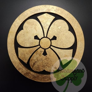 May include: A gold-colored wooden circle with a black five-petal flower design. The flower has a round center and the petals are heart-shaped. The circle is surrounded by a black border.