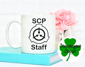 May include: White ceramic mug with a black logo that says "SCP Staff" and a black symbol with arrows pointing in different directions. The mug is sitting on a blue book. A green shamrock sticker with the text "Shenanigans Supply" is on the right side of the image.