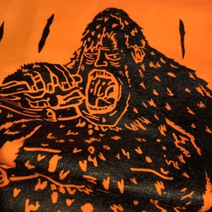 Op de afbeelding: Een zwarte en oranje grafische print van een brullende gorilla op een feloranje achtergrond.