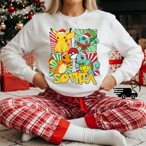 May include: White sweatshirt featuring four cartoon characters with Santa hats. The characters are Pikachu, Bulbasaur, Charmander, and Squirtle. The name "SOPHIA" is printed in yellow below the characters. The person is wearing red and white plaid pajama pants.