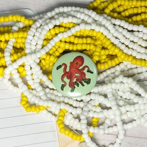 May include: A light green button with a red octopus illustration and green accents. The button is surrounded by strands of white and yellow beads. The beads are seed beads, creating a colourful and textured background.