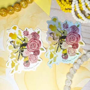 Könnte beinhalten: Zwei Aufkleber mit einem Bienen- und Rosen-Design in Rosa und Rot. Die Biene ist schwarz und gelb. Die Aufkleber haben einen weißen Rand und den Text "ShyShyShoppe Creations".