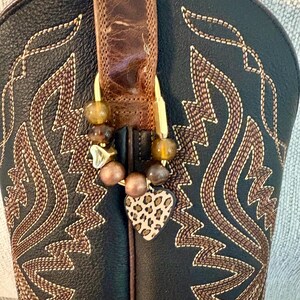 May include: A black leather cowboy boot with brown stitched patterns featuring a gold-toned metal carabiner clip attached to the pull strap. The clip is adorned with round brown beads and a flat heart-shaped charm with a tan and black leopard print.