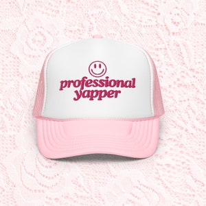 Peut inclure: Un chapeau de camionneur rose et blanc avec un smiley rose et le texte "professional yapper" en rose.