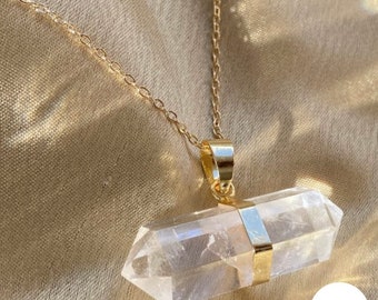 Puede incluir: Un collar de cadena dorada con un colgante de cristal de cuarzo transparente. El colgante tiene forma de un rectángulo largo y delgado con extremos puntiagudos.