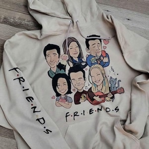 May include: A beige hoodie with a cartoon illustration of the six main characters from the TV show "Friends" on the front. The characters are wearing casual clothing and are holding various objects, including a guitar, a cup of coffee, and a cat. The text "F.R.I.E.N.D.S" is printed on the front of the hoodie, and the text "F.R.I.E.N.D.S" is printed on the left sleeve.
