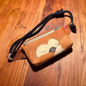 Puede incluir: Un cinturón de cuero marrón con un diseño floral cosido en la parte delantera. El cinturón tiene un cierre de cremallera y una correa de cuerda negra. El diseño floral está hecho de parches de cuero beige y marrón.