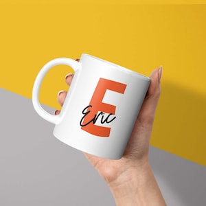 May include: White ceramic mug featuring a large orange letter "E" and the name "Eric" in black script. The mug is held against a yellow and grey background, a personalised coffee cup.