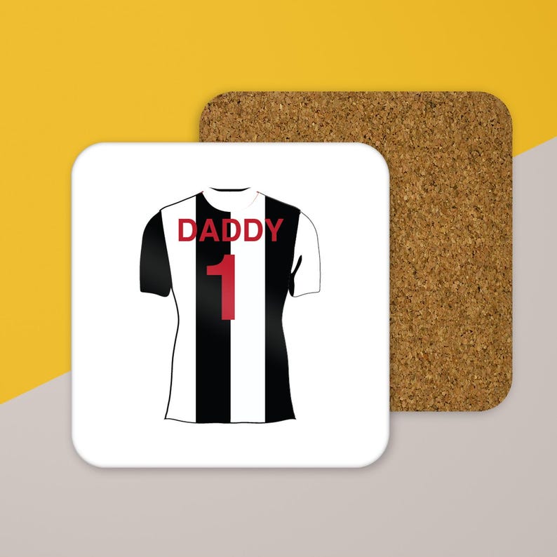 May include: A square coaster with a cork backing and a white top surface featuring a graphic of a black and white vertically striped sports jersey. The jersey has the word DADDY printed in red above the red number 1 on the front centre.