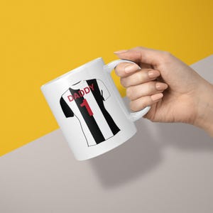 May include: A white ceramic mug featuring a graphic of a black and white striped sports jersey. The jersey design includes the red text DADDY above the red number 1. The mug has a curved handle and a smooth finish, displaying a clean and simple aesthetic.