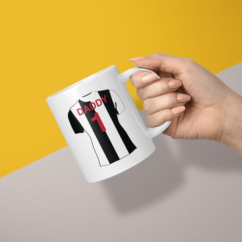 May include: A white ceramic mug featuring a graphic of a black and white striped sports jersey. The jersey design includes the red text DADDY above the red number 1. The mug has a curved handle and a smooth finish, displaying a clean and simple aesthetic.
