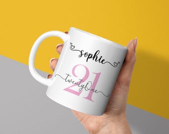 May include: White ceramic mug with a curved handle. The mug features the name "Sophie" in black script, with the number "21" in pink, and the words "Twenty One" below the number. The mug is held against a yellow and gray background.