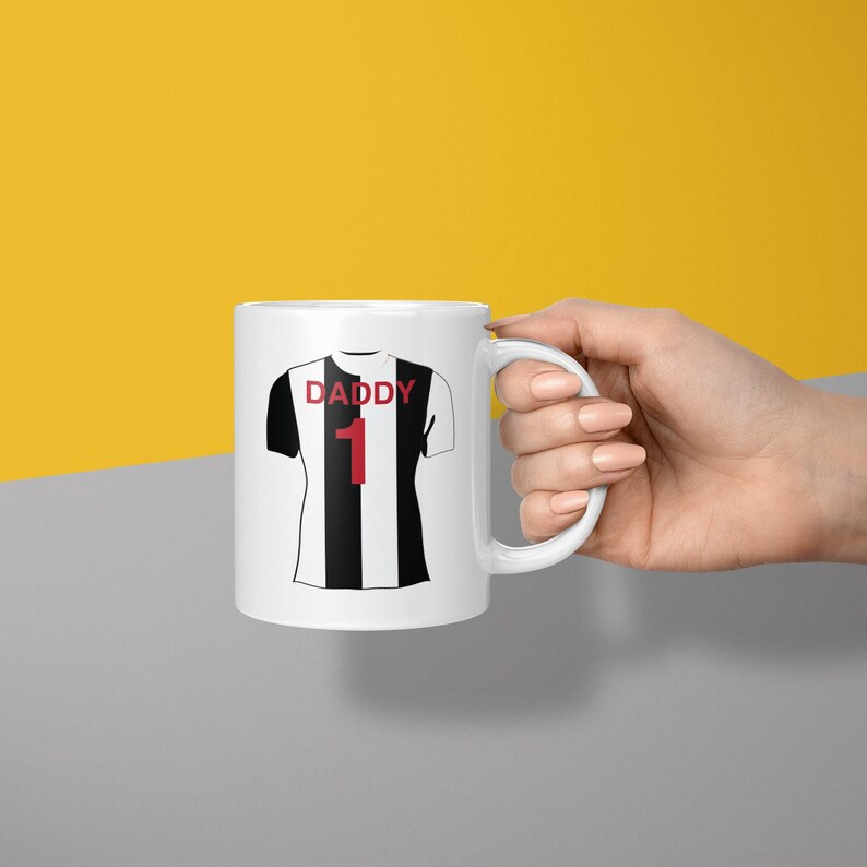May include: A white ceramic mug featuring a graphic of a black and white vertically striped sports jersey. The jersey graphic includes the red text DADDY above the red number 1. The mug has a standard handle and a smooth, glossy finish.