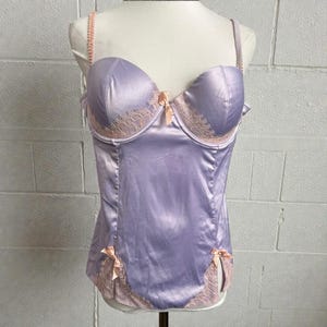 May include: A lavender satin bustier with a sweetheart neckline, featuring peach-coloured lace trim and small bows. The bustier has adjustable straps and a structured bodice, displayed on a mannequin against a white brick wall.