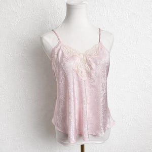 May include: A pale pink camisole top with delicate spaghetti straps and a lace trim. The top features a subtle floral design and is displayed on a mannequin. The fabric has a smooth, satin-like texture.