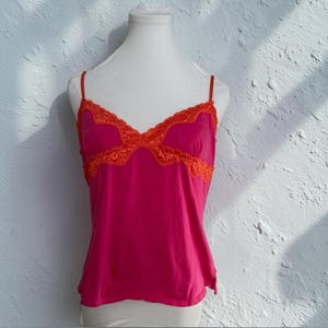 May include: A bright pink camisole with thin spaghetti straps and orange lace trim. The lace trim is around the neckline and the top of the camisole. The camisole is displayed on a white mannequin.