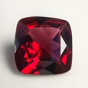 May include: A faceted, cushion-cut gemstone with a deep red hue. The gem's surface reflects light, creating bright red flashes within the darker, burgundy body. The square shape has rounded corners, and the gem is set against a white background.
